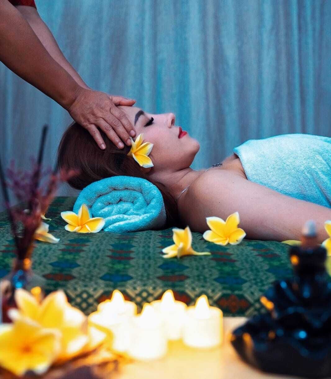 Een persoon ontvangt een massage, met een handdoek om het hoofd, omringd door gele bloemen en kaarsen op een patroonrijk oppervlak.