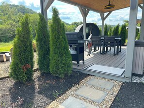 A covered outdoor patio with a grill, seating, and potted trees.