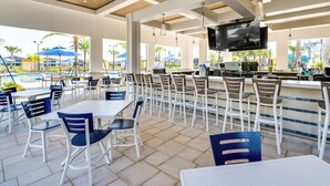 An outdoor dining area with white tables and chairs, a bar with a flat-screen TV, and a pool in the background.