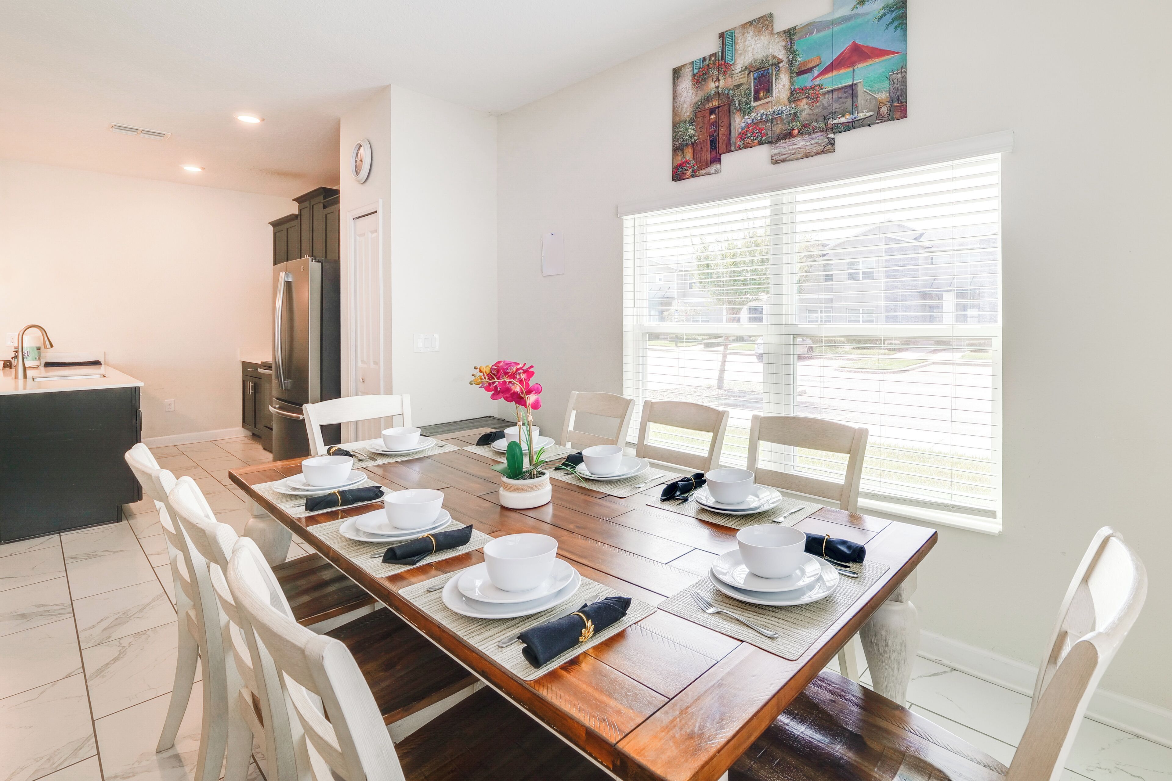 A dining area with a wooden table set for four, white chairs, a vase with flowers, and a painting on the wall.
