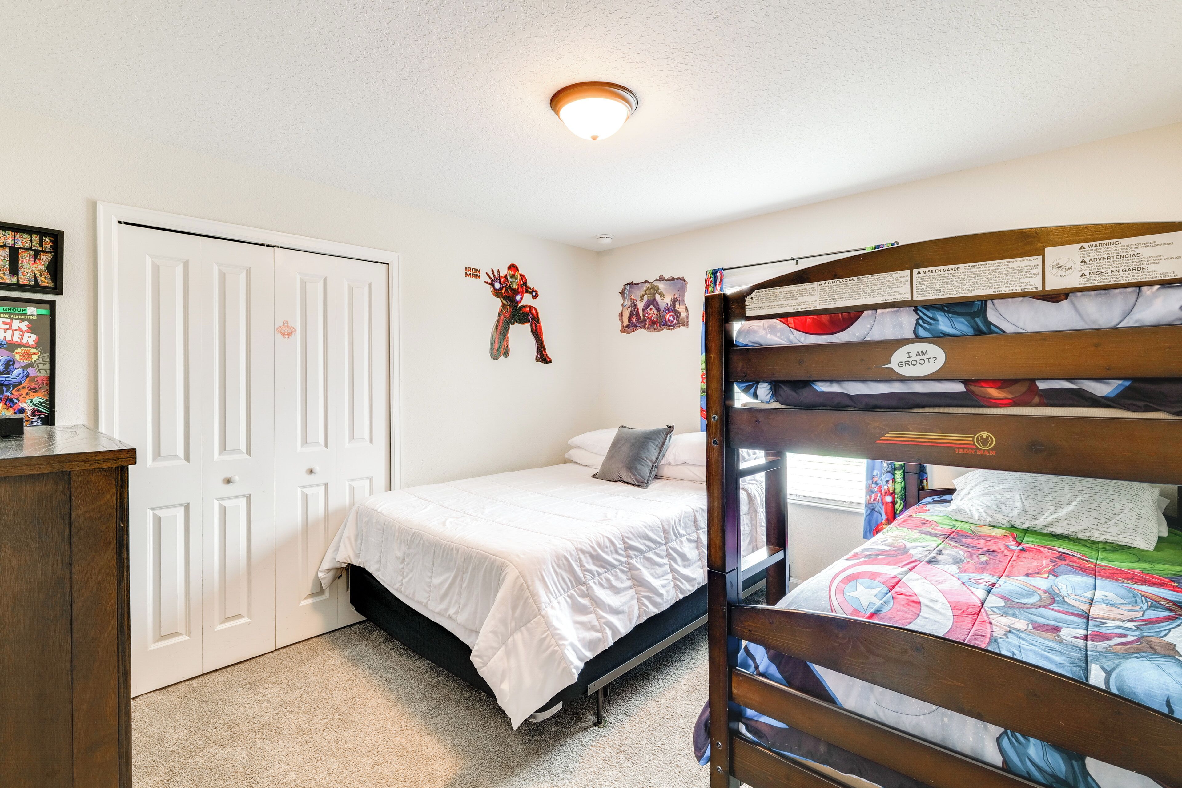 A bedroom with a bunk bed, a single bed, a door, and a wall with superhero-themed decorations.
