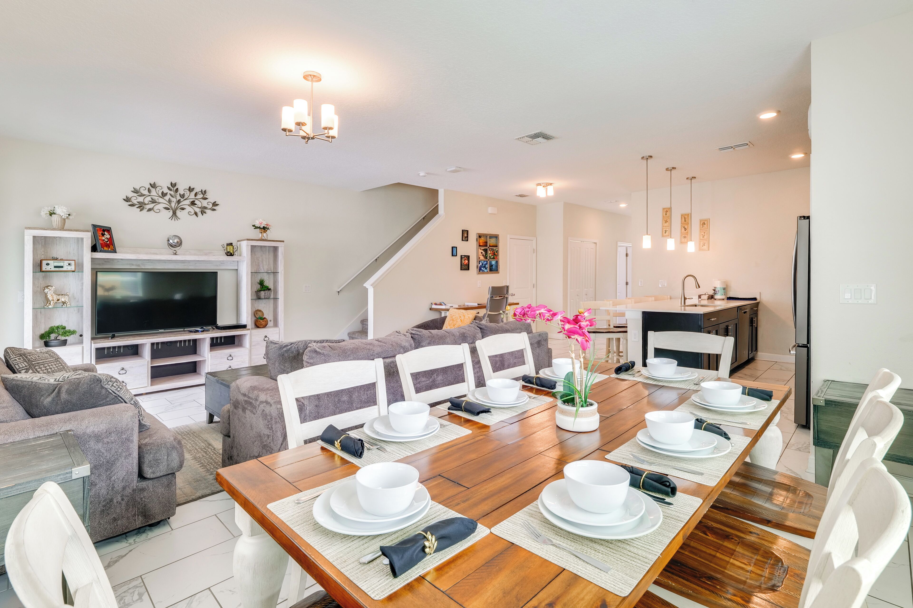 Una sala moderna con un televisor grande, sofás grises, una mesa de comedor preparada para seis personas y una cocina al fondo.