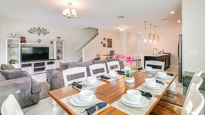 A modern living room with a large TV, grey sofas, a dining table set for six, and a kitchen in the background.