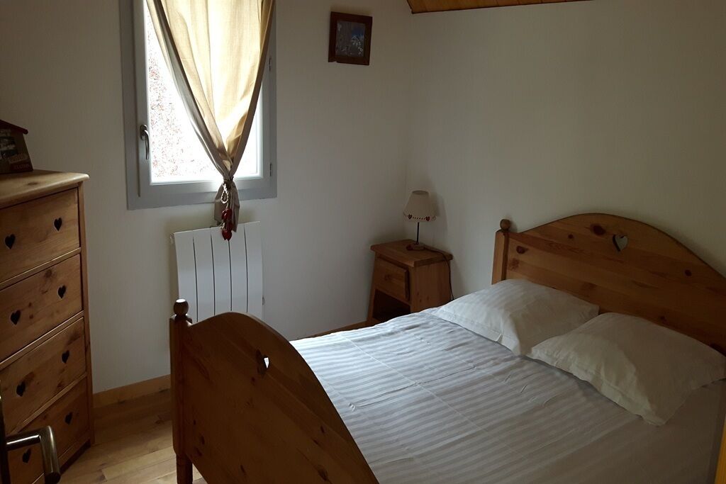 Quarto com cama de madeira, cômoda de madeira com puxadores em formato de coração, mesa de cabeceira com abajur e janela com cortinas.