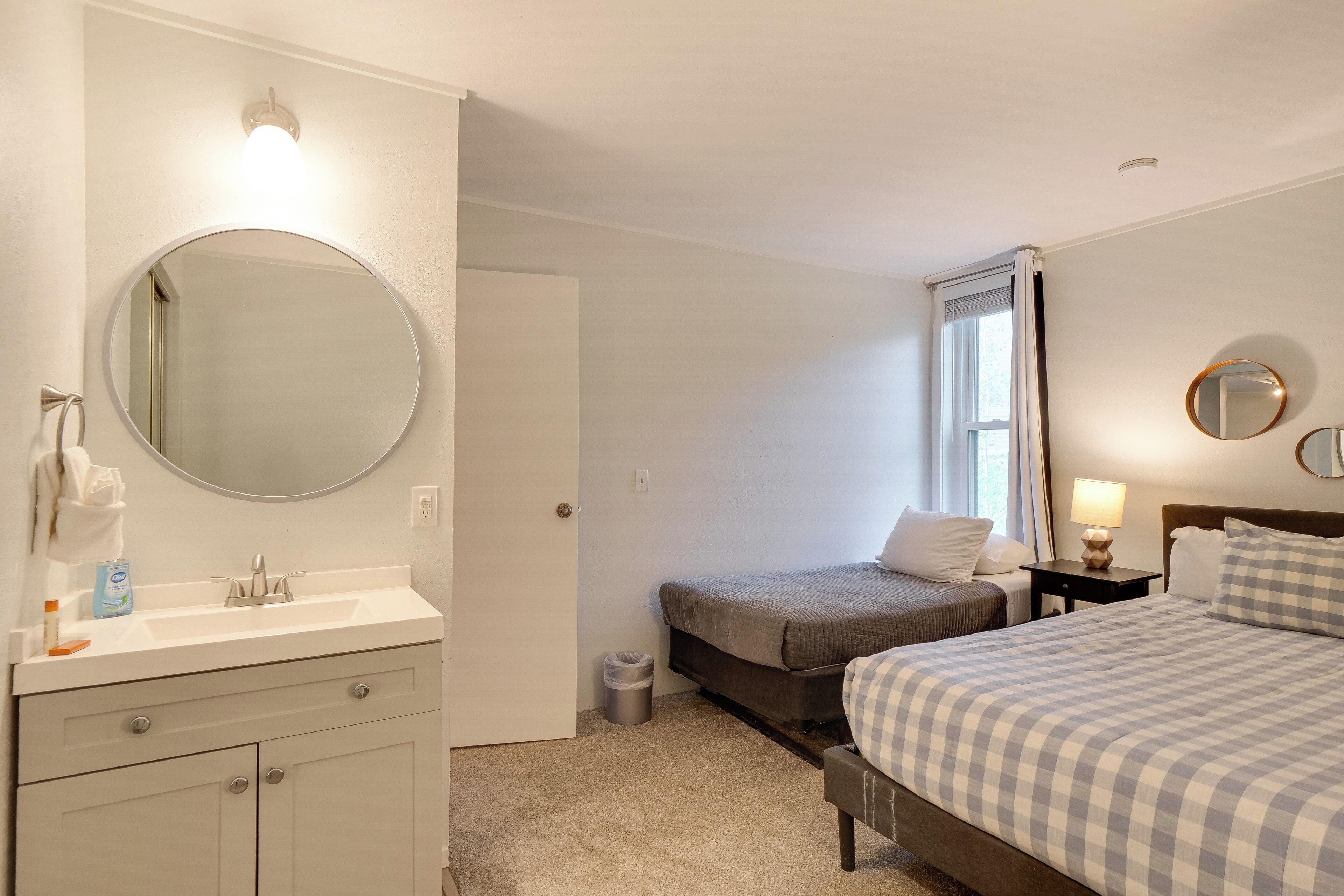 A hotel room with a bed, a bedside table with a lamp, a small bedside table with a lamp, a round mirror above the sink, a towel rack, and a trash can.