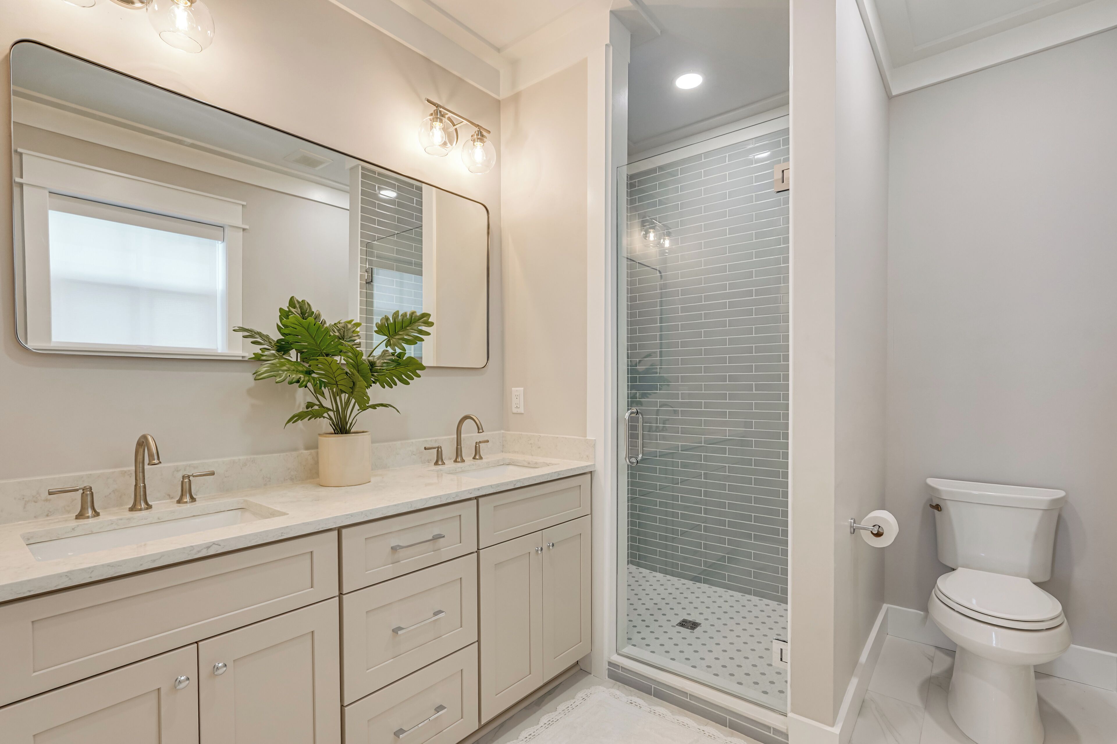 A modern bathroom with a large mirror, two sinks, a potted plant, and a glass shower enclosure.