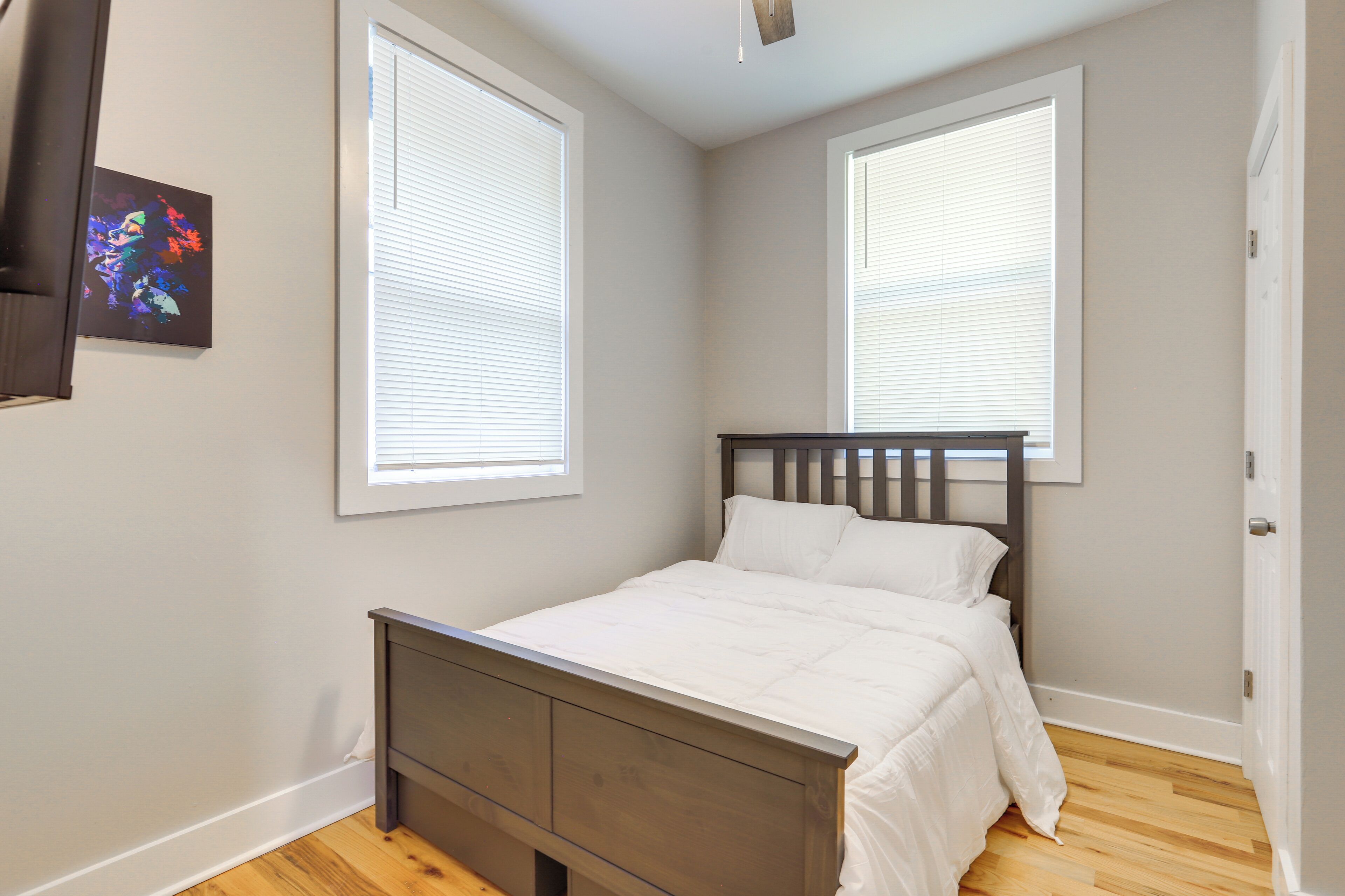Un dormitorio con una cama de madera, ropa de cama blanca y dos ventanas con persianas.
