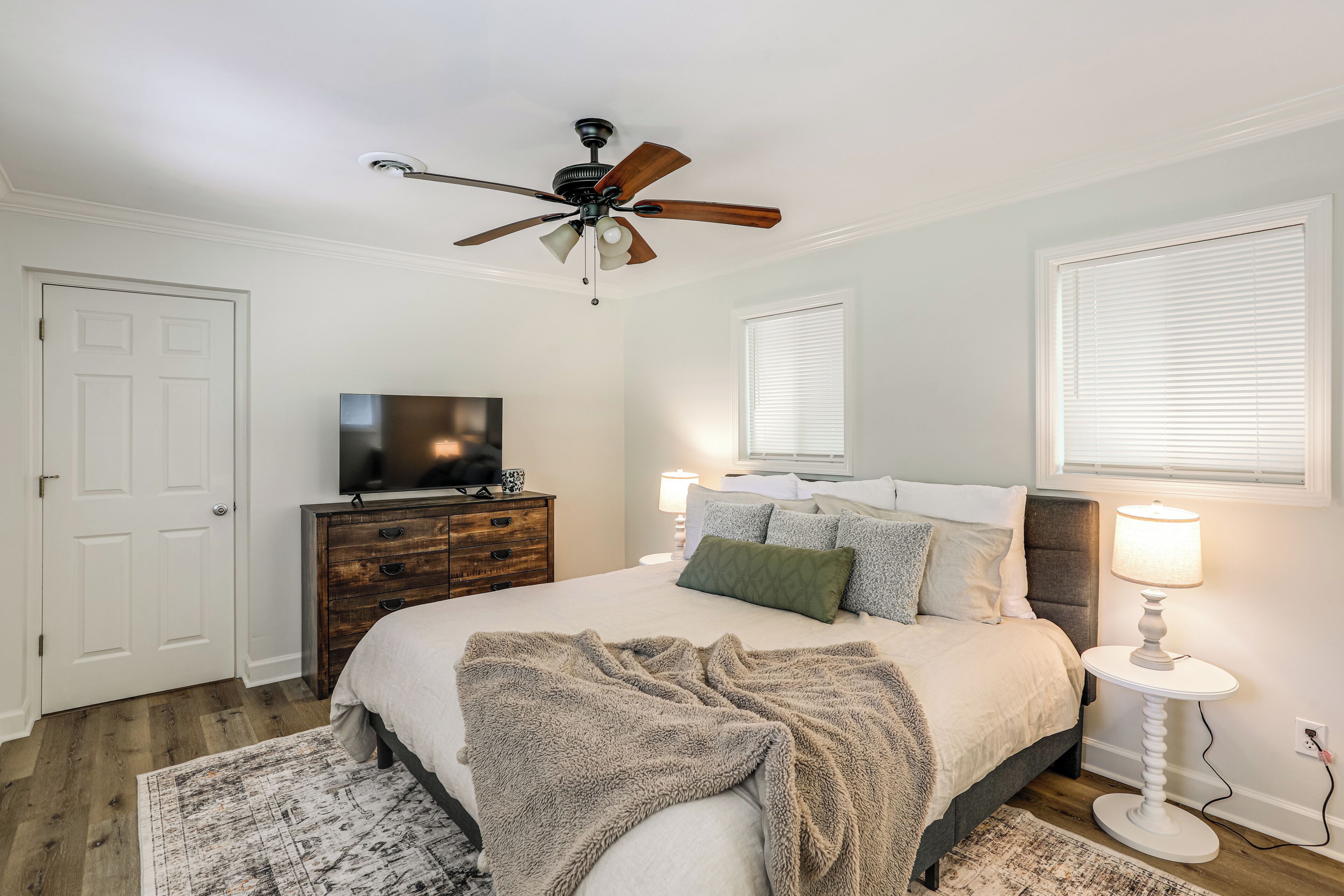 A bedroom with a bed, a ceiling fan, a television, a bedside table with a lamp, and a window with blinds.