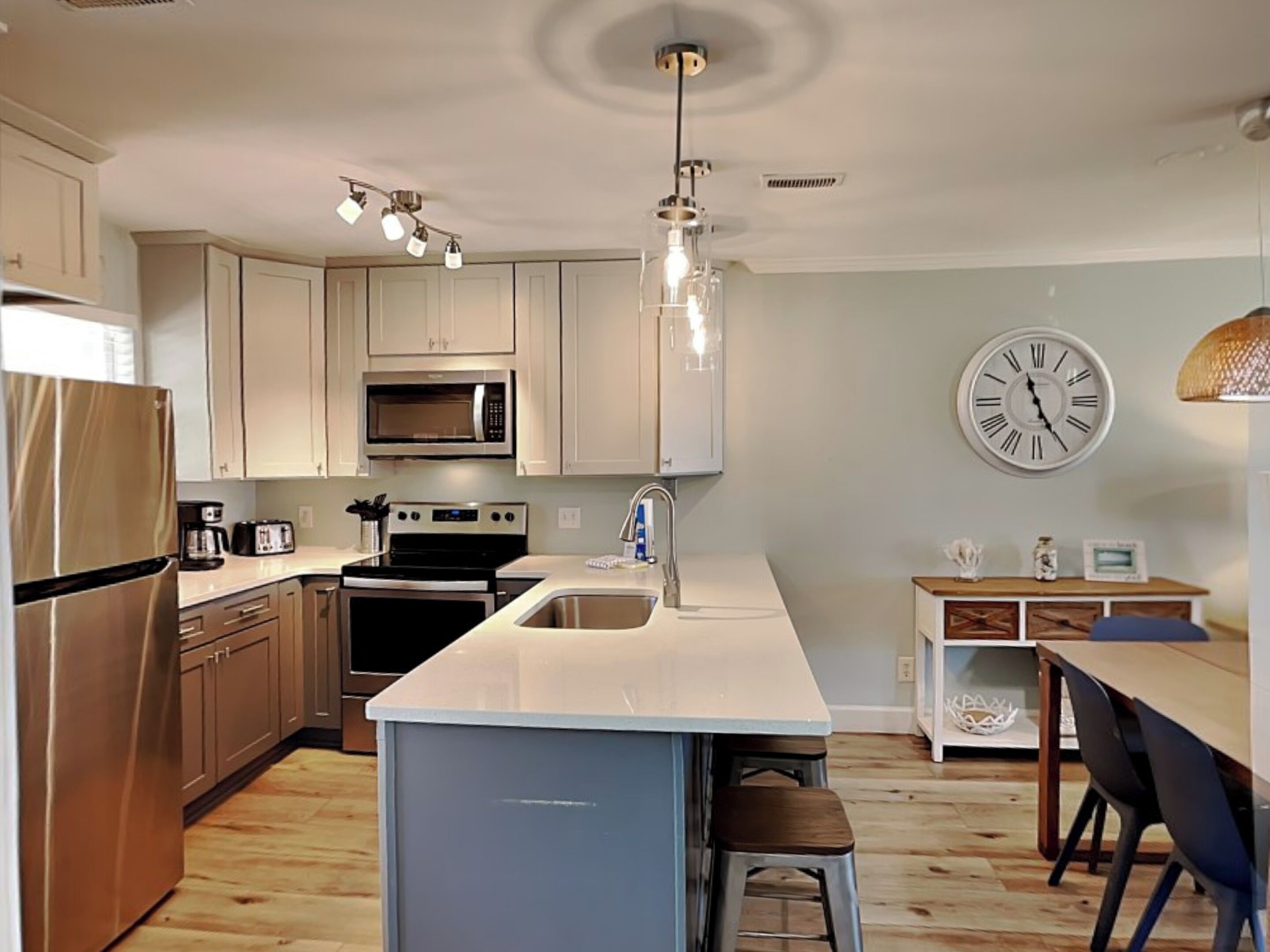 A modern kitchen with stainless steel appliances, a central island with stools, and a dining area with a clock on the wall.