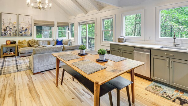 A spacious kitchen with a wooden table, grey cabinets, and large windows offering a view of the outdoors.