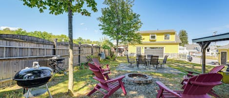 A backyard with a barbecue grill, red Adirondack chairs, a fire pit, and a yellow house in the background.
