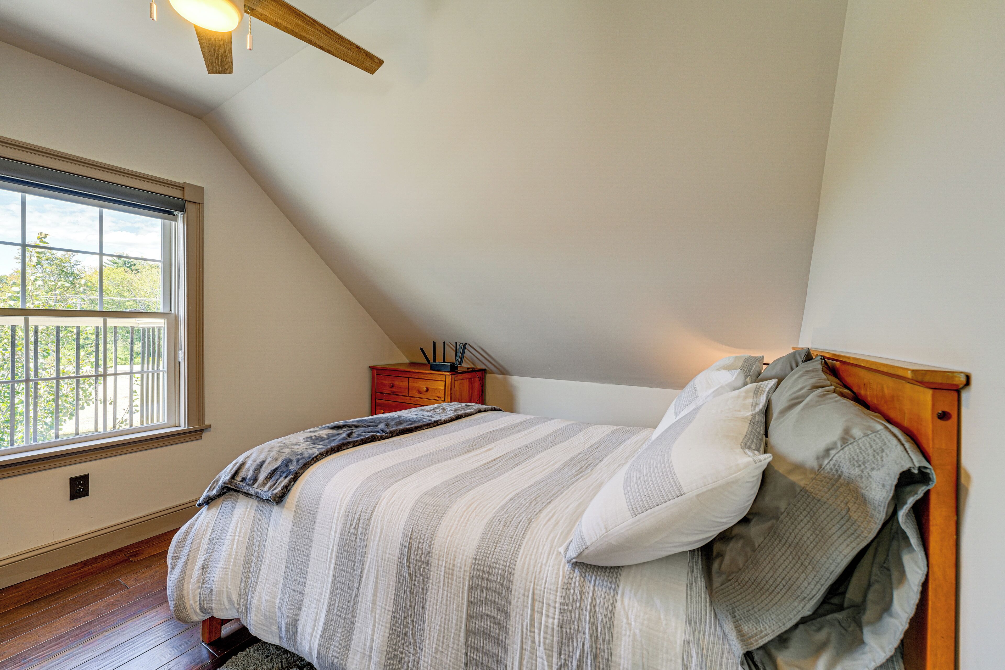 A bedroom with a sloped ceiling, a wooden headboard, a bed with striped bedding, a nightstand with a lamp, and a window with a view of trees.