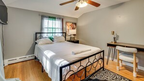 Ein Schlafzimmer mit einem Bett, einem Schreibtisch, einem Stuhl, einem Deckenventilator und einem Fenster mit Vorhängen.