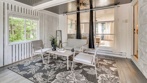 Ein modernes Wohnzimmer mit einem weißen Sofa, einem Couchtisch und einem gemusterten Teppich. Ein großes Fenster mit Jalousien, eine Tür mit Vorhang und ein Deckenventilator sind ebenfalls vorhanden.