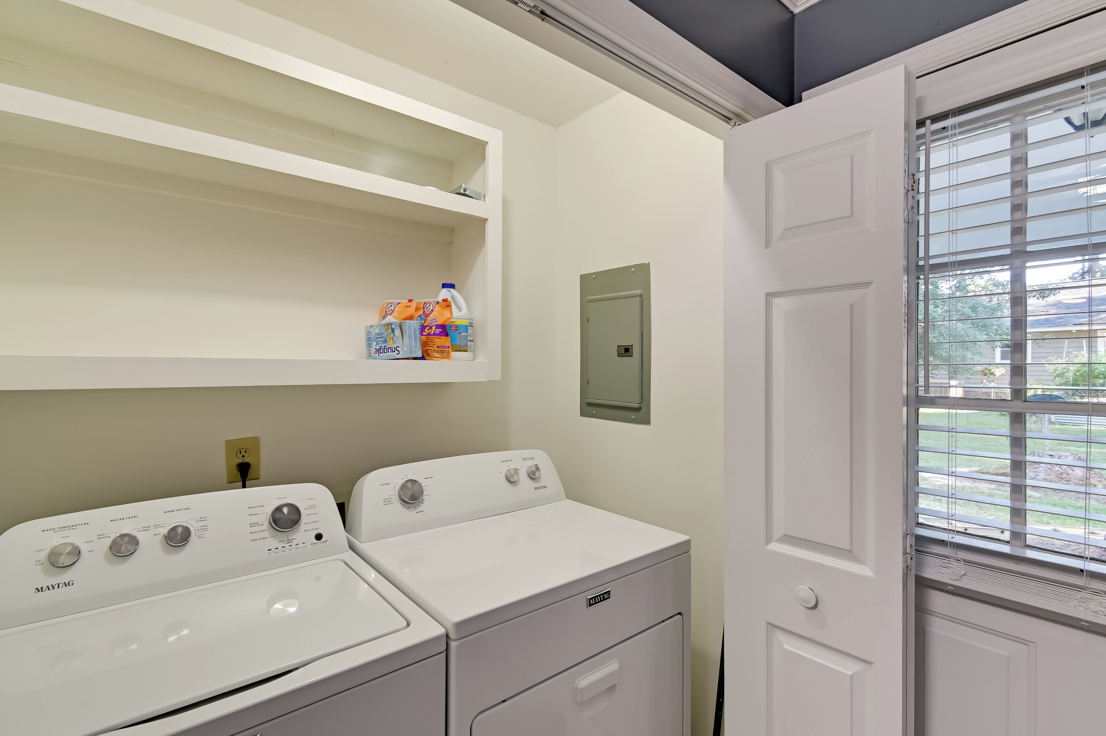 Un lavadero con dos lavadoras y secadoras Maytag, un estante con productos de limpieza y una puerta que da al exterior.