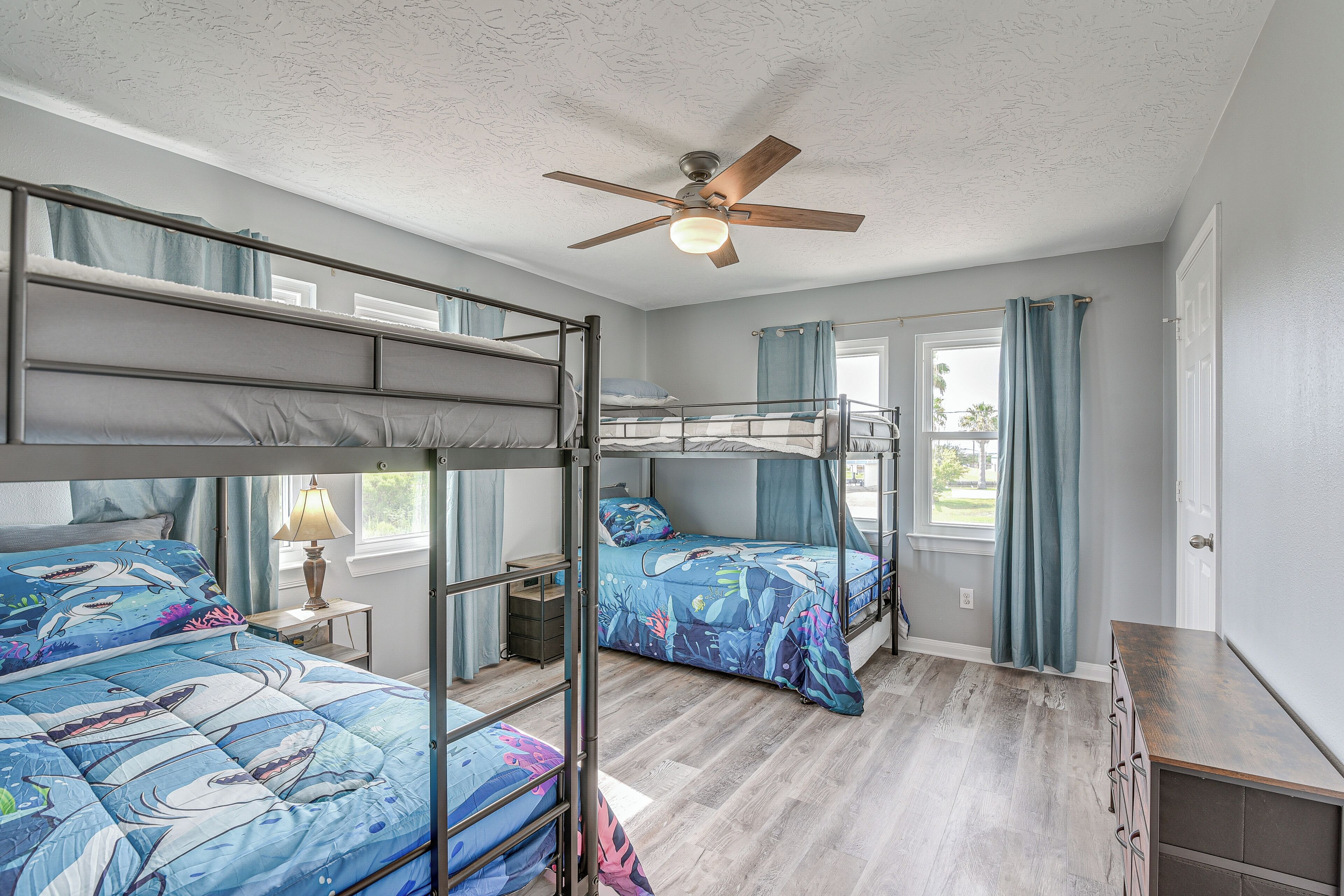 A bunk bed room with a ceiling fan, a window with curtains, and a bedside table with a lamp.