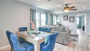 A dining area with a wooden table and blue upholstered chairs, a ceiling fan, and a view into a living room with a sofa and a rug.