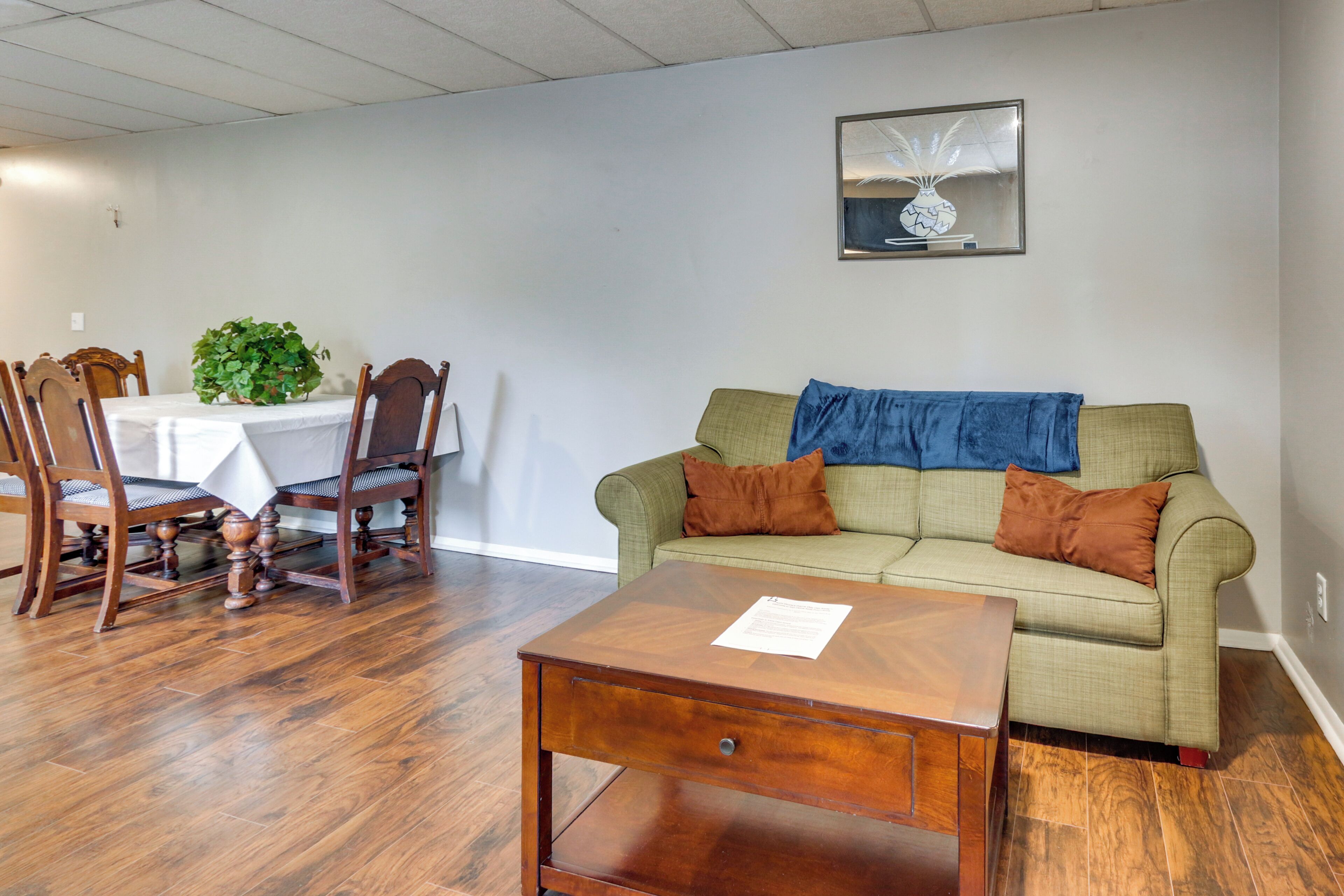 A living room with a dining table, wooden chairs, a green sofa with cushions, a coffee table, and a framed picture on the wall.