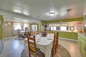 A dining area with a table set for four, surrounded by chairs, a floral centerpiece, and a kitchen area with green cabinets and a window with floral curtains. - Hike & Fish: Rustic, Timeless Mckee Home w/ Yard (McKee)