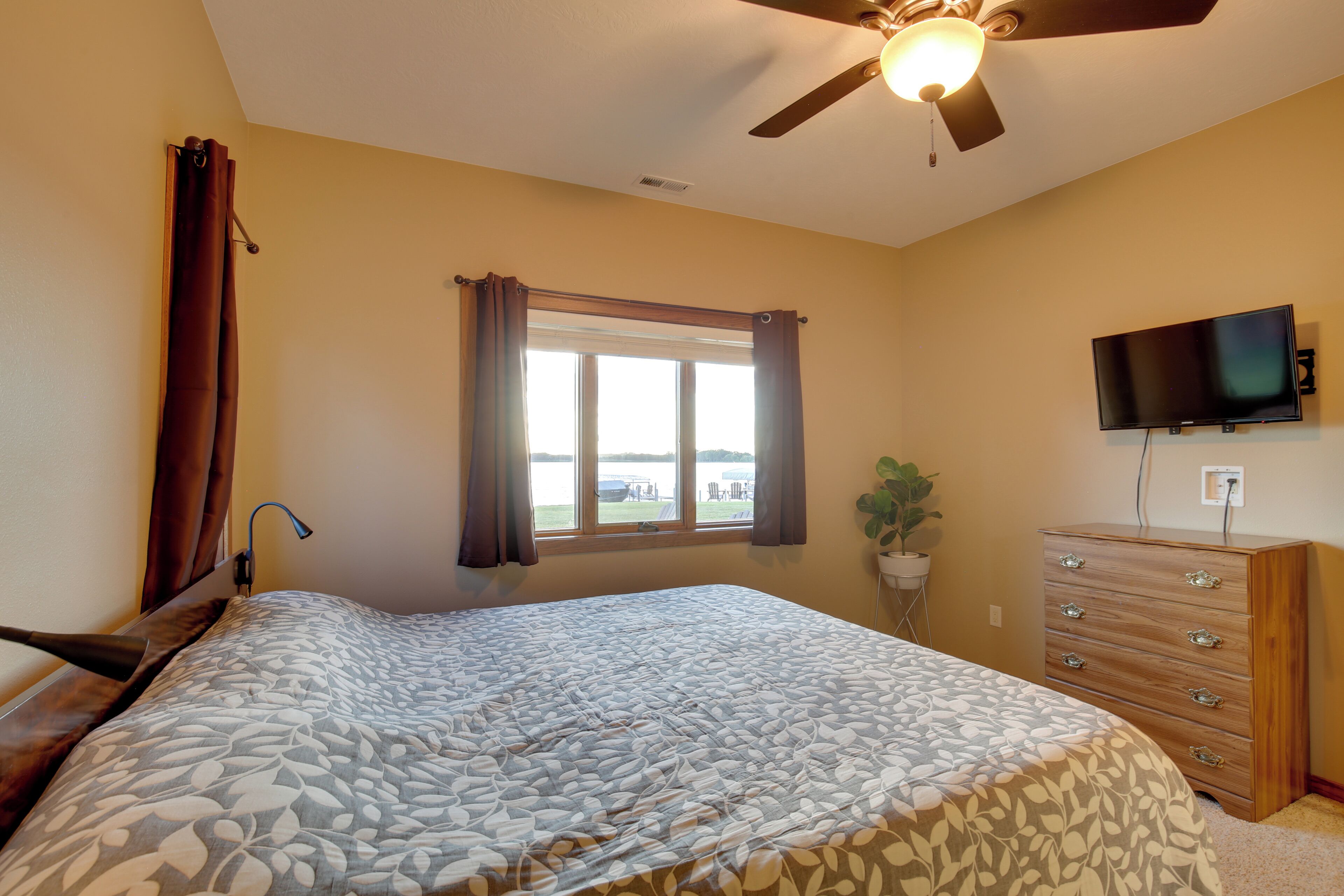 Un dormitorio con cama, ventilador de techo, ventana con cortinas, cómoda y televisor.