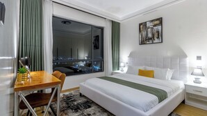 Habitación de hotel con una cama grande, un escritorio, una silla, una ventana con cortinas, un cuadro en la pared y una mesita de noche con lámpara.