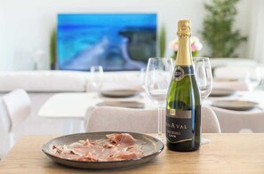 Un tavolo da pranzo con una bottiglia di cava, un piatto di cibo e bicchieri, con una televisione che mostra un panorama.