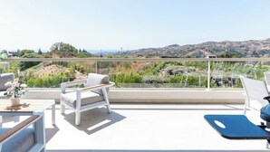 En balkon med udsigt over et bølgende landskab, en hvid stol, en blå træningsmåtte og et lille bord med en plante.