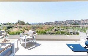 Un balcone con vista su un paesaggio collinare, una sedia bianca, un tappetino da ginnastica blu e un tavolino con una pianta.