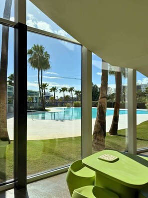 Una habitación moderna con ventanas amplias, piscina, palmeras y una mesa verde.