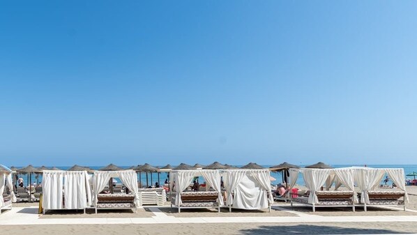 Un hotel frente a la playa con varias camas blancas cubiertas por toldos, dispuestas en filas, un cielo azul despejado y el océano al fondo.