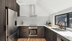 A modern kitchen with dark wood cabinets, a stainless steel refrigerator, a built-in oven, and a window offering a view of the countryside. - Central Escape - A modern townhouse with mountain and lake views in the heart of Queenstown. (Queenstown)