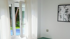 A bedroom with a bed, a window with curtains, a radiator, a small table, and a framed picture on the wall. - Arena Surf Hotel (Somo)