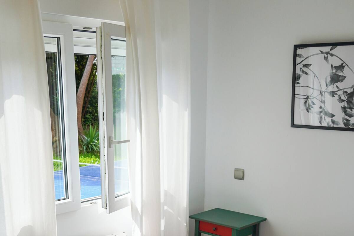 A bedroom with a bed, a window with curtains, a radiator, a small table, and a framed picture on the wall.