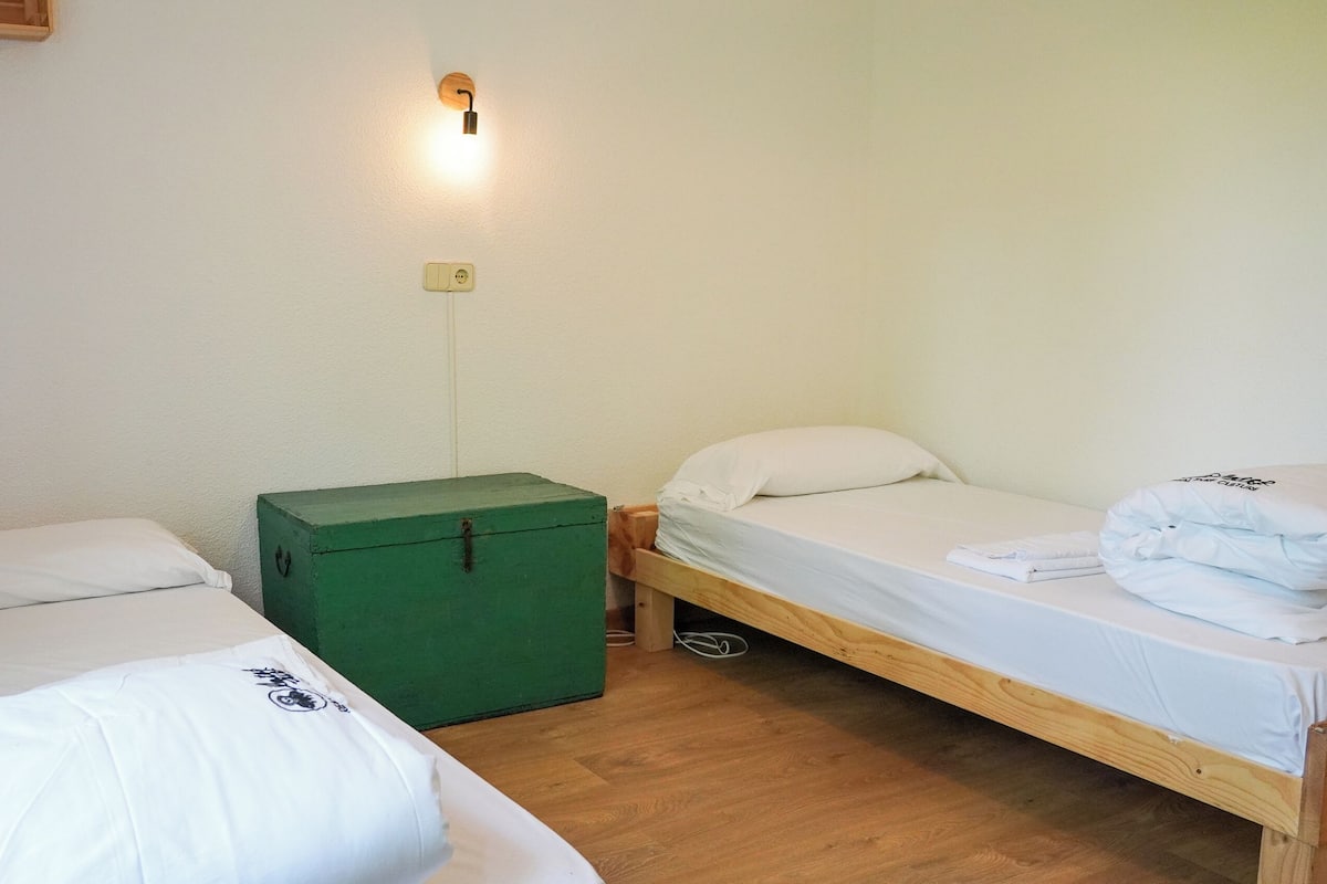 A small, simple room with two single beds, a green storage box, a wall-mounted light, and a power outlet.