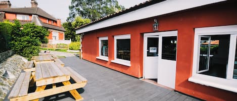 Un edificio rojo con ventanas blancas y una puerta, una terraza de madera con mesas de picnic, y un muro de piedra al fondo.