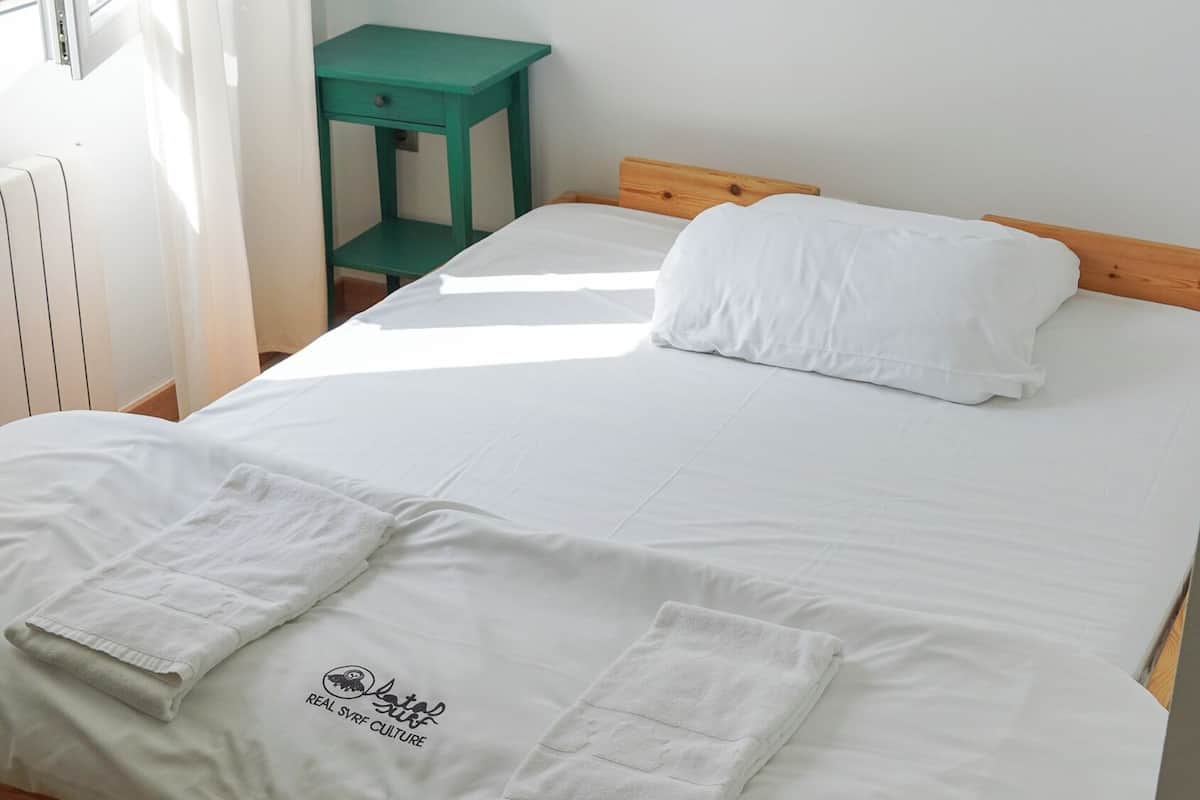A neatly made bed with white linens and towels, a wooden headboard, and a small green table beside it.