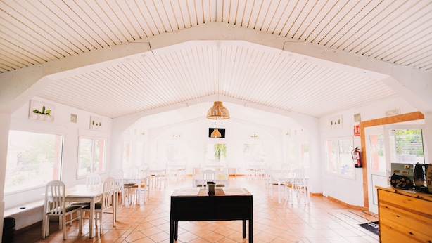 A spacious room with a high ceiling, white walls, and a tiled floor. There is a table with chairs in the center, and a wooden cabinet on the right. - Arena Surf Hotel (Somo)