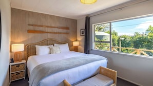 A bedroom with a large bed, bedside lamps, a wicker bench, a view of greenery, and a wooden wall panel.