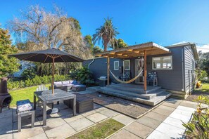 A patio with a table and chairs, a hammock, and a covered seating area. - Beach Bliss – Steps to Onetangi (Waiheke Island)
