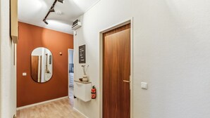 Un pasillo con suelo de madera, una alfombra negra, una puerta de madera, una pared blanca, un espejo, un armario blanco, un extintor y una lámpara en el techo.