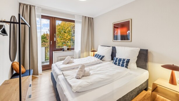 Habitación de hotel con una cama grande, una ventana con cortinas, un radiador, un perchero y un cuadro en la pared.
