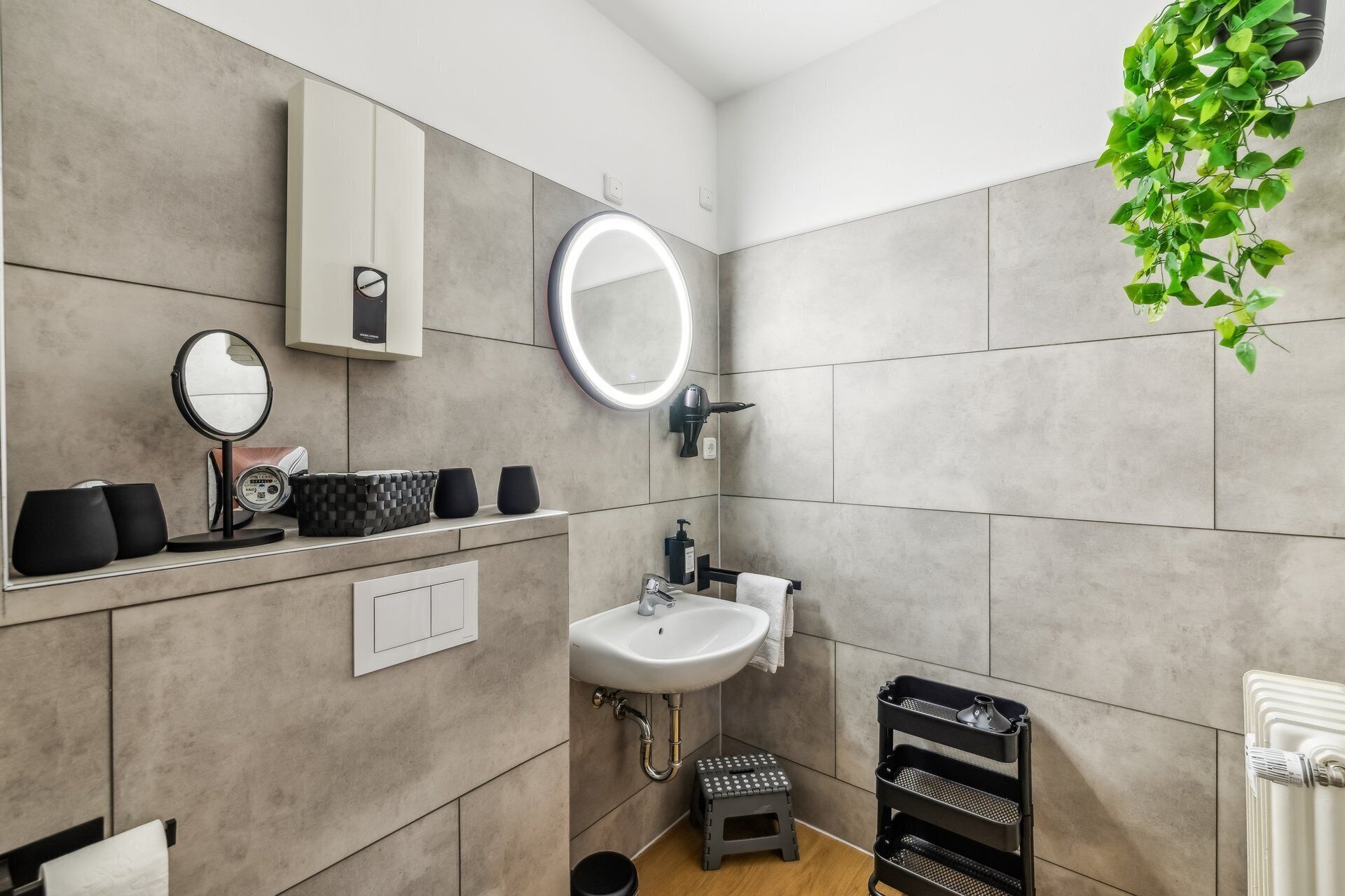 Un baño moderno con baldosas grises, un espejo redondo, un lavamanos, un taburete pequeño y un estante negro con artículos de aseo.