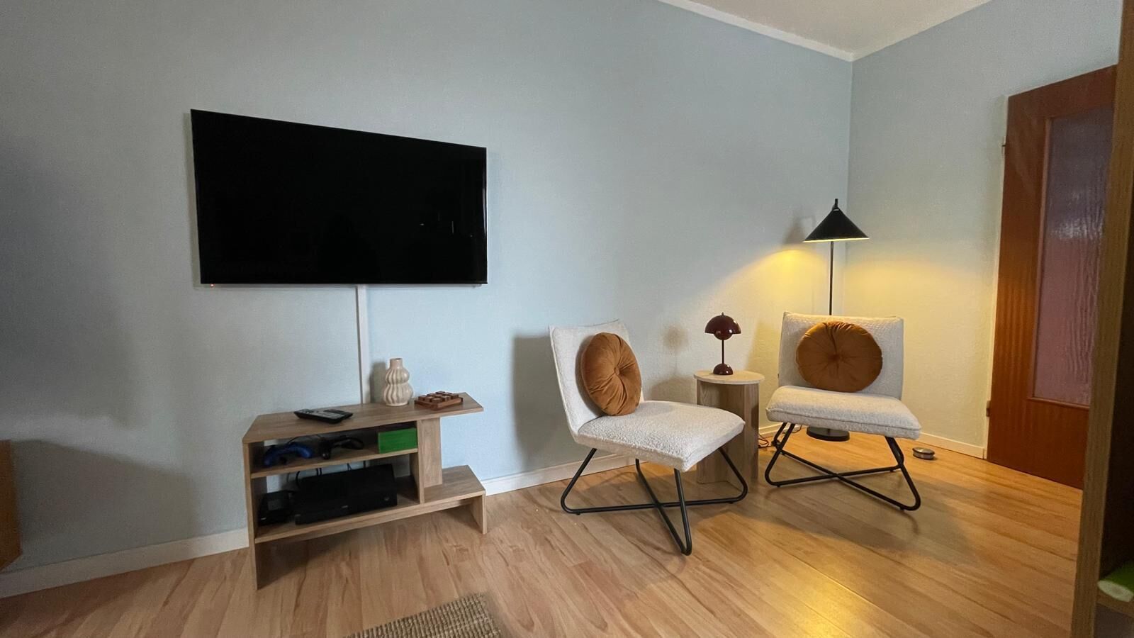 A living room with a flat-screen TV mounted on the wall, a wooden entertainment unit with electronic devices, two modern chairs, and a floor lamp.