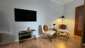 A living room with a flat-screen TV mounted on the wall, a wooden entertainment unit with electronic devices, two modern chairs, and a floor lamp.