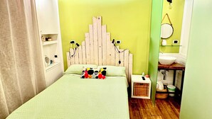 Un dormitorio con pared verde, cabecera de madera, cama con colcha estampada, mesita de noche, espejo y una ventana pequeña.