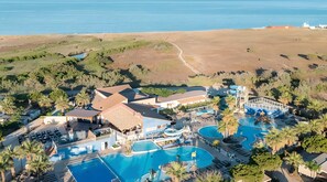 Vista aèria d'un complex turístic amb diverses piscines, una platja i un fons de paisatge montanyós.