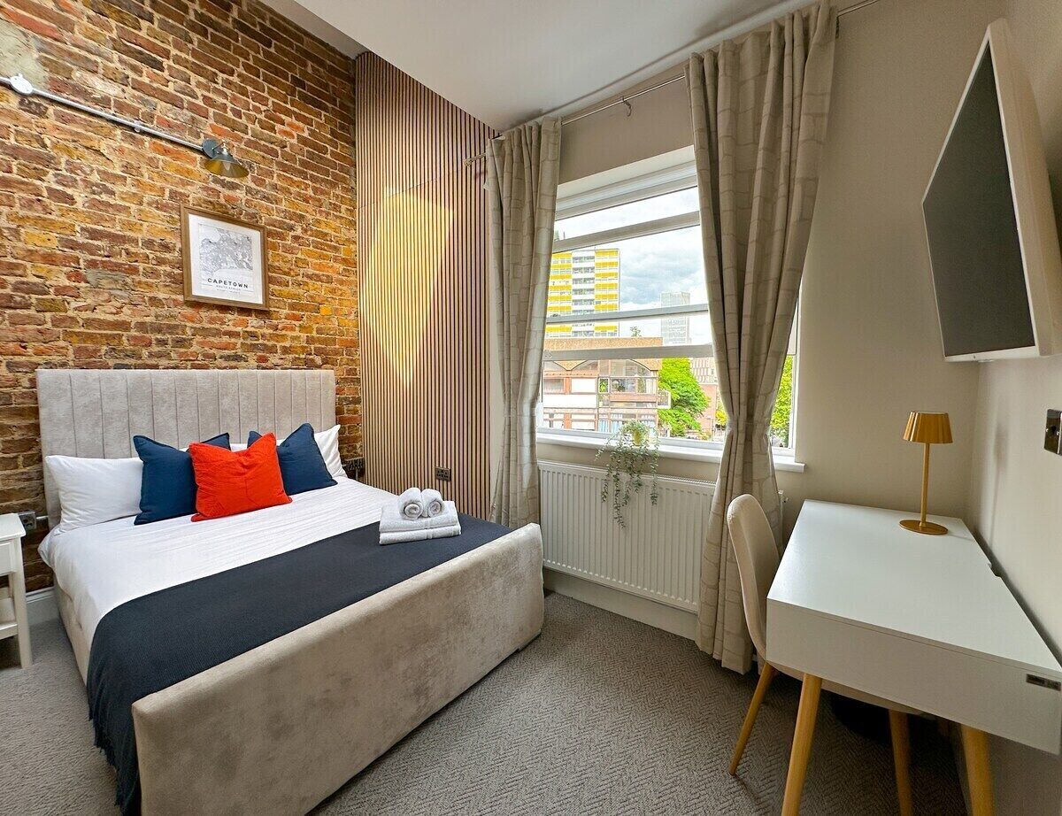 A hotel room with a brick wall, a bed with a headboard, a desk, and a window with curtains.
