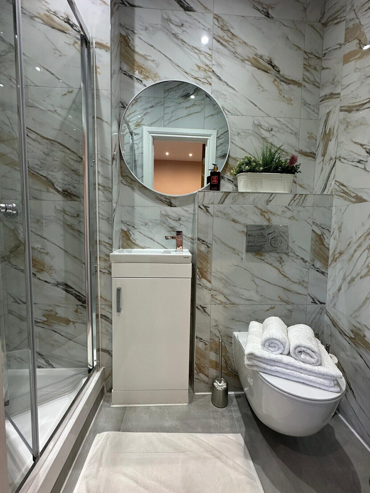 A bathroom with a glass shower enclosure, a white sink, a round mirror, a toilet, and a towel rack.