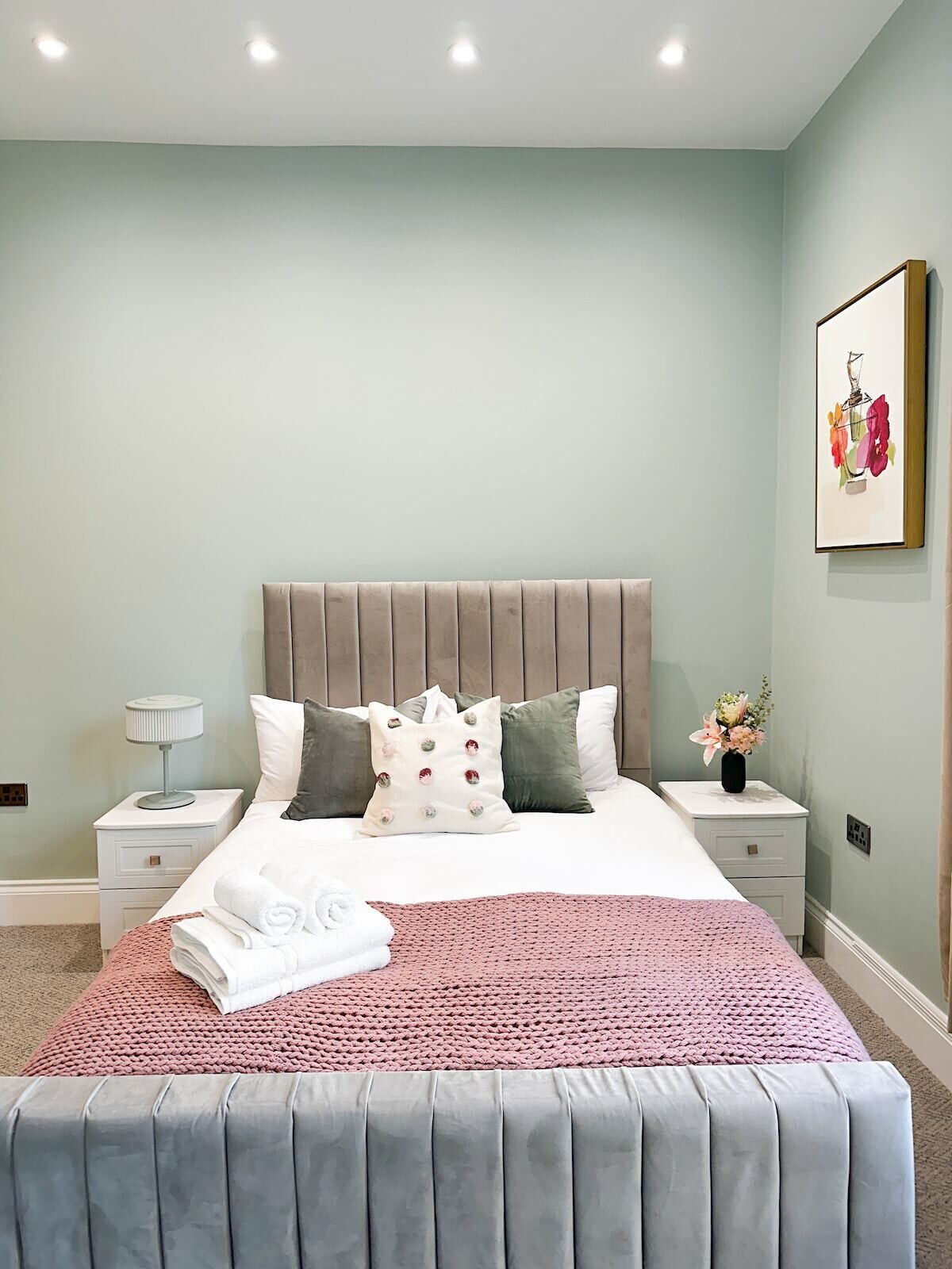 Ein ordentlich eingerichtetes Schlafzimmer mit einem großen Bett, einem Nachttisch mit Lampe und einem gerahmten Bild an der Wand.