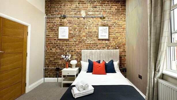 A bedroom with a brick wall, a bed with a headboard, a nightstand, a window with curtains, and a door. - Farringdon Rooms (London)